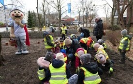 przedszkolaki sadzą kwiaty, w tle maskotka miasta Marianek