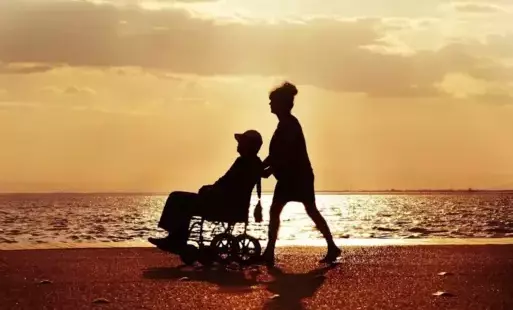 na tle zachącego słońca na plaży kobieta pcha po piasku wózek z osobą z niepełnosprawnością 